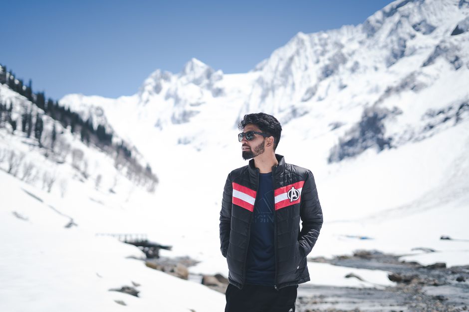 A man standing in a snowy mountain landscape wearing sunglasses and a stylish jacket, embodying winter fashion.