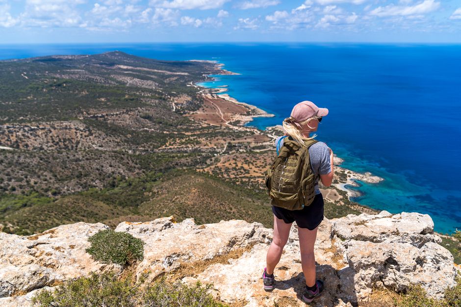Back view full body of anonymous female hiker with backpack standing on rocky cliff while enjoying view of blue sea during trip